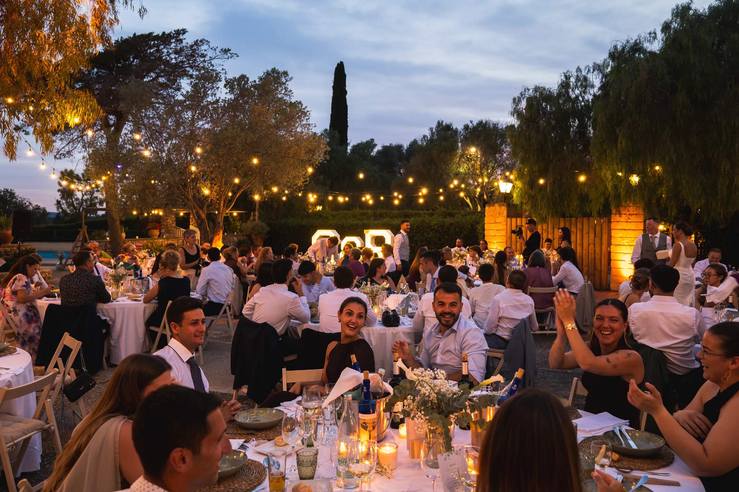 Banquete de boda de noche en el jardín, con invitados bajo guirnaldas de luces